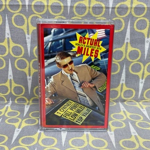 May include: A red cassette tape case with a photo of a man in a brown jacket and sunglasses holding a cigar. The text on the case reads "Actual Miles Henley's Greatest Hits". There is a sign on the car that reads "1 Owner Full Power Low Miles Top Value".
