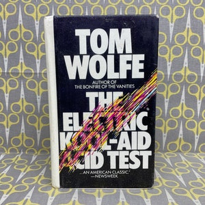 May include: A white book cover with black text that reads "TOM WOLFE" and "THE ELECTRIC KOOL-AID ACID TEST". The author is listed as "AUTHOR OF THE BONFIRE OF THE VANITIES". The cover has a colorful, abstract design with streaks of yellow, pink, and blue.