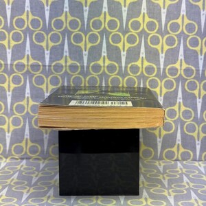 May include: A book with a dark cover and yellowed pages is displayed on a black stand. The background features a repeating pattern of yellow and white scissors on a gray fabric. The book's title and author are visible.