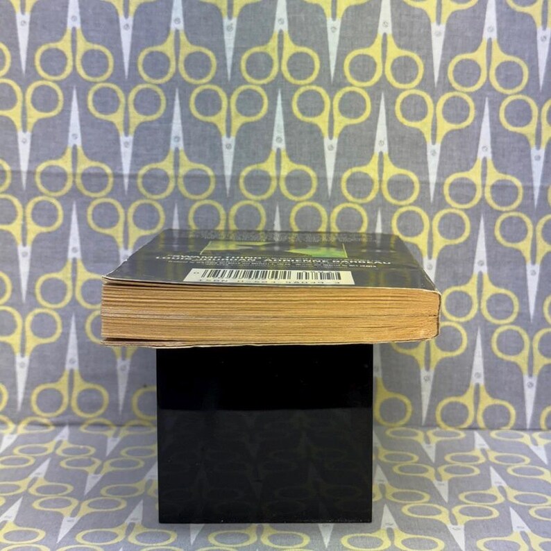 May include: A book with a dark cover and yellowed pages is displayed on a black stand. The background features a repeating pattern of yellow and white scissors on a gray fabric. The book's title and author are visible.