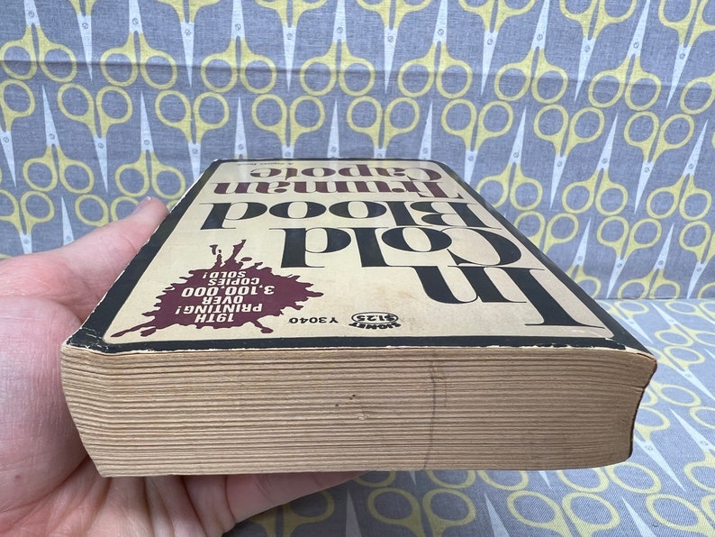 May include: A close-up of the spine of a paperback book titled "In Cold Blood" by Truman Capote. The book is worn and has a brown cover with black text. The text on the spine indicates that this is the 19th printing and over 3,100,000 copies have been sold.