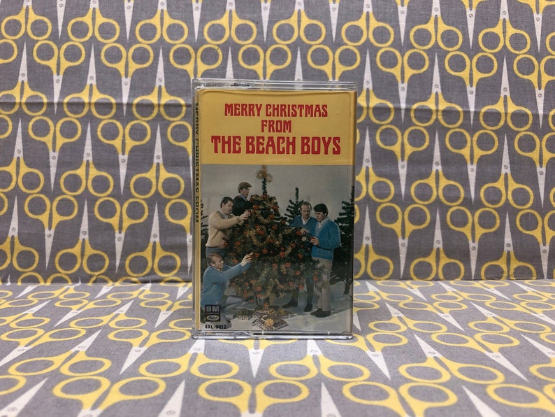 May include: A vintage cassette tape with a yellow and red label. The label reads "Merry Christmas From The Beach Boys". The image shows the band members standing around a Christmas tree.