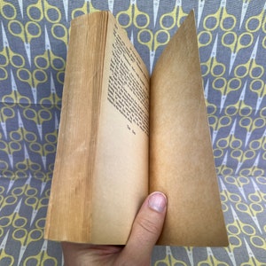 May include: An open book with yellowed pages and a brown cover. The book is lying open on a gray and yellow fabric with a pattern of scissors.