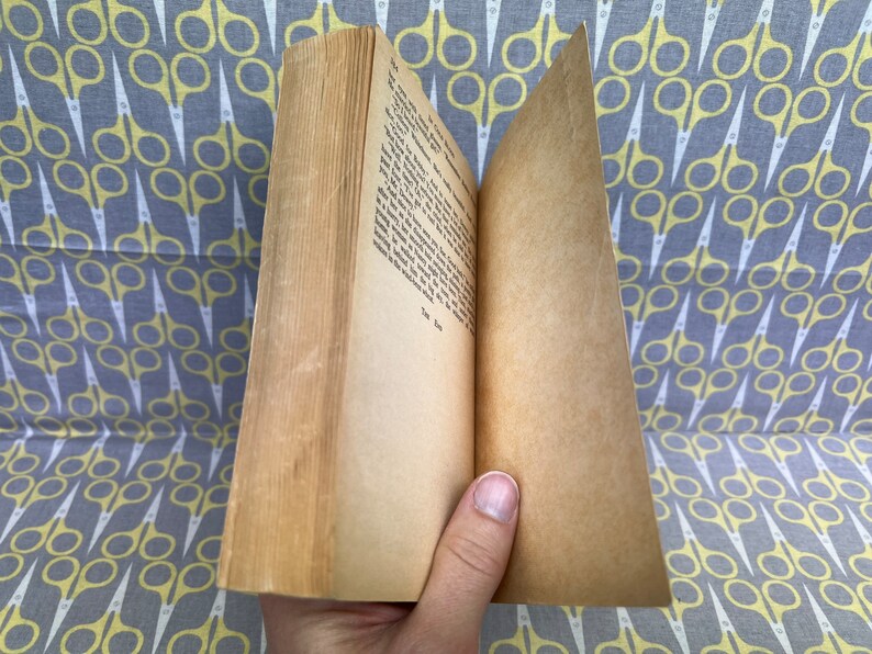 May include: An open book with yellowed pages and a brown cover. The book is lying open on a gray and yellow fabric with a pattern of scissors.