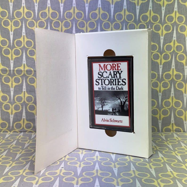 Puede incluir: Un libro de tapa dura titulado "MORE SCARY STORIES to Tell in the Dark" de Alvin Schwartz, exhibido dentro de una caja de cart&oacute;n blanca. La portada del libro presenta una ilustraci&oacute;n oscura y espeluznante de una escena forestal.