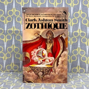 Op de afbeelding: Een vintage paperback boekomslag met een rode, oranje en gele illustratie van een vrouw in een rode mantel die een gloeiende bol vasthoudt. De titel van het boek is "ZOTHIQUE" door Clark Ashton Smith.
