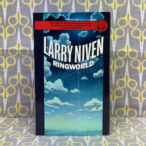 May include: A paperback book cover with a blue background featuring a large white cloud and a ring-shaped world with a bright light in the center. The title "RINGWORLD" is in white letters and the author "LARRY NIVEN" is in white letters. The book cover also includes a small circle with the text "SF" in white letters.