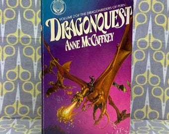 May include: A vintage paperback book titled "Dragonquest" by Anne McCaffrey. The cover features a golden dragon breathing fire, with a rider on its back, set against a purple and blue background. The book is part of the "Dragonriders of Pern" series.
