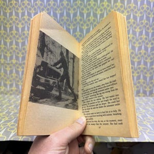 May include: An open vintage book with aged, yellowed pages. A black and white illustration depicts a person pushing a vehicle. The text on the pages is in English. The book is held open, revealing the text and illustration.