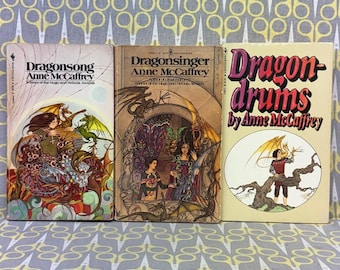 以下が含まれることがあります： アン・マキャフリイの3冊のヴィンテージペーパーバック。書籍のタイトルは「Dragonsong」、「Dragonsinger」、「Dragondrums」です。表紙には、ドラゴンや人物が描かれたファンタジーイラストが鮮やかな色で描かれています。本は模様のある表面に表示されています。