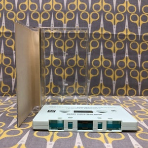 May include: A white cassette tape with the text "Merry Christmas From" printed on the front. The tape is in a clear plastic case.