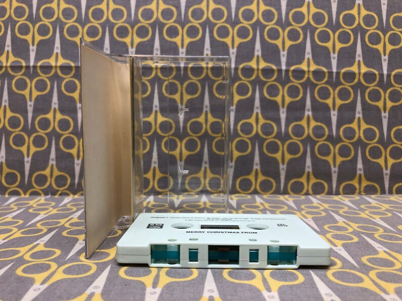 May include: A white cassette tape with the text "Merry Christmas From" printed on the front. The tape is in a clear plastic case.