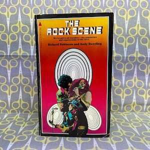 May include: A vintage paperback book titled "The Rock Scene" with a red, orange, and black cover featuring a psychedelic design of concentric circles and a photo of a band performing. The book's subtitle reads "We've got it all together - the superstars and supersounds of the 70's!" and the authors are listed as Richard Robinson and Andy Zwerling.