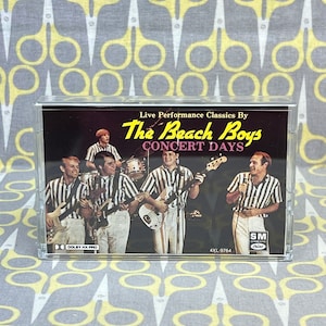 May include: A cassette tape case with a black background featuring a photo of The Beach Boys band members. The text on the case reads "Live Performance Classics By The Beach Boys Concert Days" and "4XL-9764".