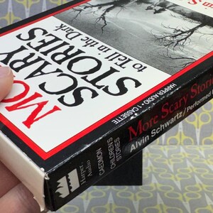 Puede incluir: Caja de cinta de cassette vintage para "More Scary Stories to Tell in the Dark". La portada presenta el t&iacute;tulo en letras grandes y en negrita con un borde rojo. La caja del casete es en blanco y negro con el nombre del autor y el logotipo del editor.