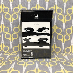 Puede incluir: Una cinta de casete negra con una etiqueta blanca. La etiqueta tiene el texto "U2 BOY" en letras blancas y un diseño abstracto en blanco y negro. El texto "U2 - BOY" también está impreso en la etiqueta. El texto "Each side contains complete album" está impreso debajo del diseño. El texto "90040-4" está impreso debajo del texto.