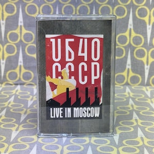 Puede incluir: Un casete vintage con un gráfico rojo y blanco que presenta el texto "UB40 CCCP LIVE IN MOSCOW". La obra de arte representa una figura estilizada y elementos industriales. El casete está dentro de una caja de plástico transparente.