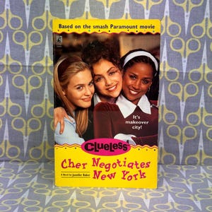 Op de afbeelding: Een paperback boek getiteld "Clueless Cher Negotiates New York" met een gele omslag. De omslag toont drie lachende vrouwen en de tekst "Based on the smash Paramount movie". Het boek is een roman van Jennifer Baker.