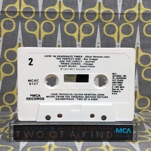 May include: A white cassette tape with the title "Two of a Kind" and the names John Travolta and Olivia Newton-John printed on the front. The cassette tape is labeled with the song titles "Livin' in Desperate Times", "The Perfect One", "Ask the Lonely", "Prima Donna", and "Night Music".