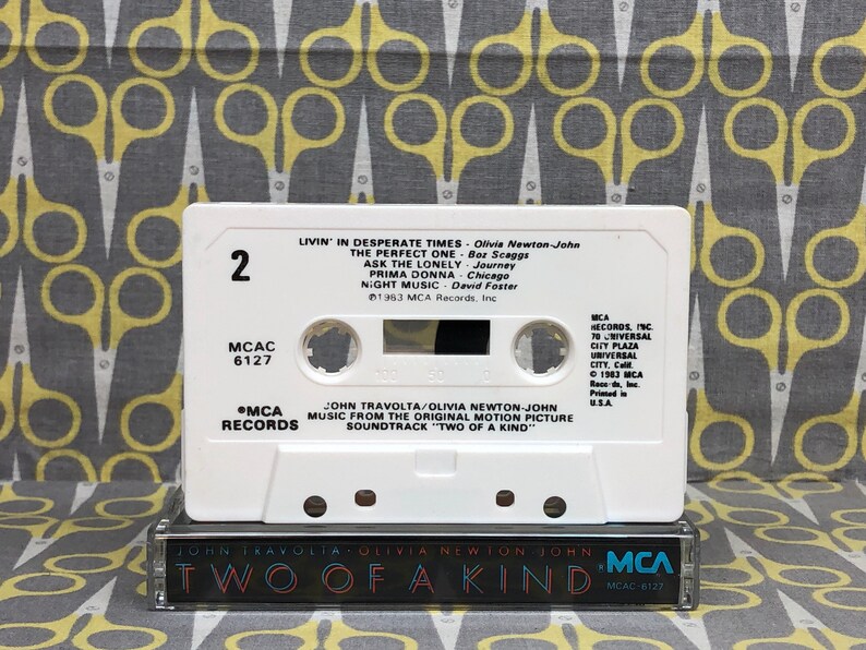 May include: A white cassette tape with the title "Two of a Kind" and the names John Travolta and Olivia Newton-John printed on the front. The cassette tape is labeled with the song titles "Livin' in Desperate Times", "The Perfect One", "Ask the Lonely", "Prima Donna", and "Night Music".