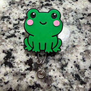 May include: A green frog-shaped badge reel with a smiling face and pink cheeks. The badge reel has a clear plastic retractable cord and a metal clip for attaching to clothing or accessories.