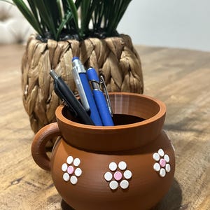 May include: A brown ceramic pen holder with a handle and three white and pink flower designs. The pen holder contains several pens in black and blue. A woven basket with green plants is in the background.