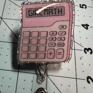 May include: A pink calculator-shaped badge reel with the text "GIRLMATH" displayed on the screen. The badge reel has a clear plastic retractable cord and a silver metal clip. The edges of the badge reel are outlined with glitter.