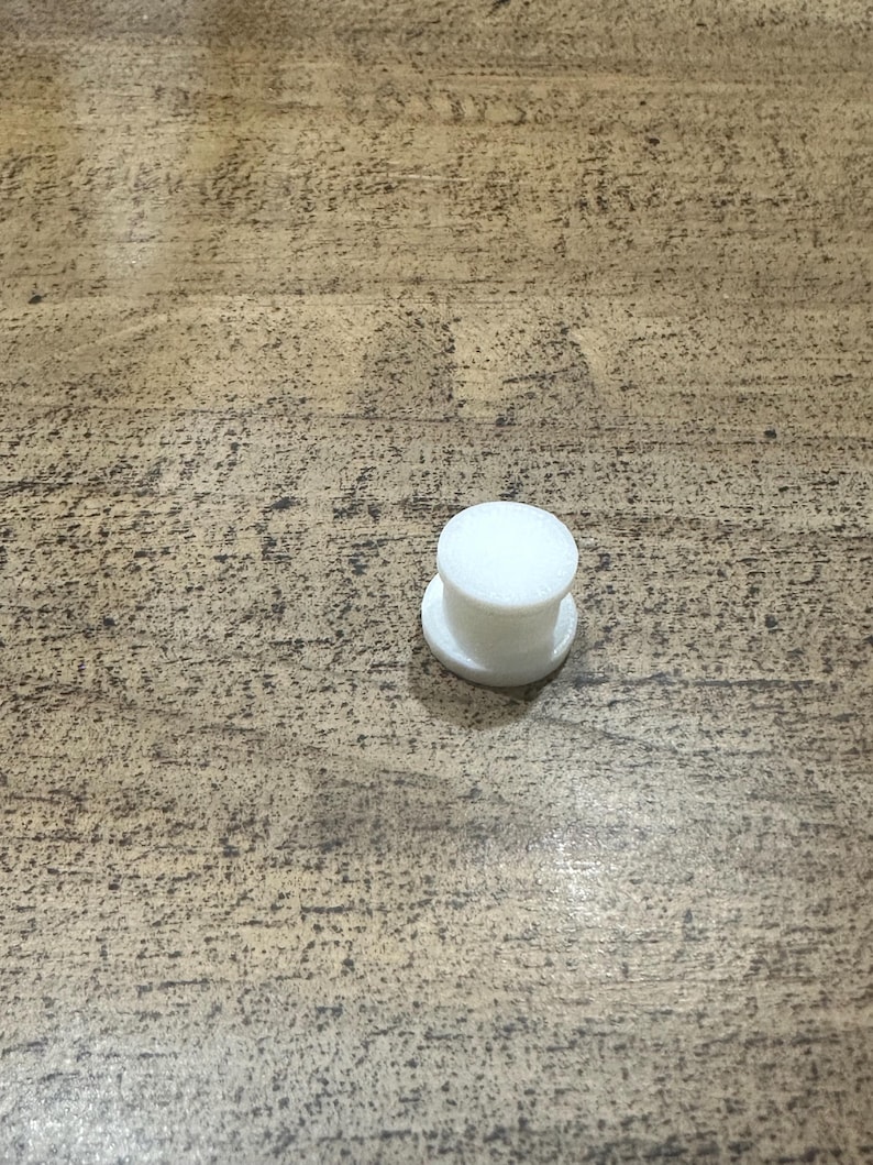 May include: A small, white, cylindrical object with a flat base and a rounded top. The object appears to be made of a solid, opaque material and is sitting on a wooden surface.