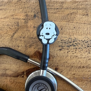 Puede incluir: Un estetoscopio negro con un dije de Snoopy adjunto. El dije es blanco con contornos negros y presenta la cara de Snoopy. El estetoscopio tiene la marca "Littmann" impresa en el diafragma.
