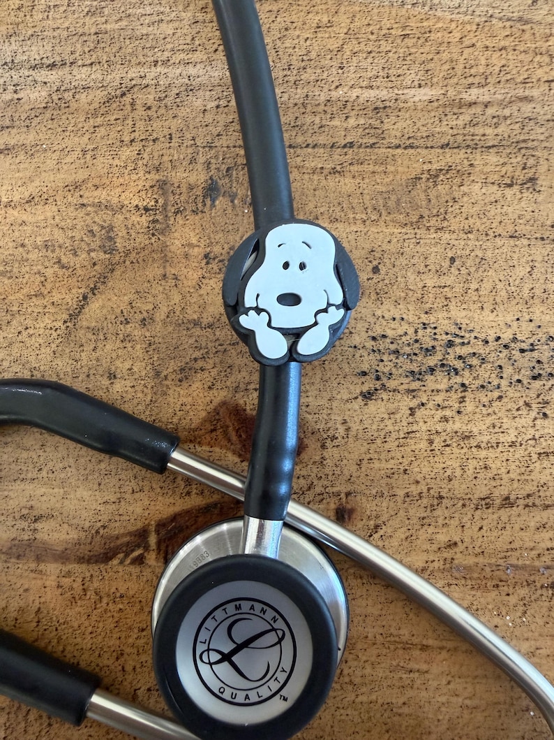 Puede incluir: Un estetoscopio negro con un dije de Snoopy adjunto. El dije es blanco con contornos negros y presenta la cara de Snoopy. El estetoscopio tiene la marca "Littmann" impresa en el diafragma.