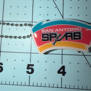 May include: A San Antonio Spurs keychain with a team logo. The keychain features a rectangular design with the team name and logo in black, red, yellow, and blue. It is attached to a silver ball chain.