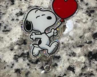 May include: A white and black cartoon dog holding a red heart-shaped balloon. The dog is on a clear background with glitter. The image is a badge holder.