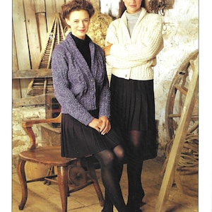 May include: A vintage fashion advertisement featuring two women modeling knitwear. One wears a purple cable-knit cardigan, the other a cream-colored cardigan. Both are paired with black skirts and tights. The Sirdar logo is at the bottom.
