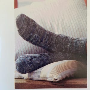 May include: A pair of blue and gray knitted socks with a cable knit pattern. The socks are resting on striped pillows. The text "Men's Socks by Martin Storey" is visible at the bottom of the image.