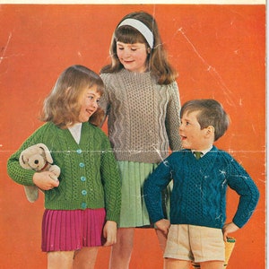May include: A vintage magazine cover showing three children in Aran knitwear. A girl wears a green cardigan, another a beige sweater, and a boy a blue sweater. The cover reads "Aran Knittings" and "sunbeam", with a chest size range.