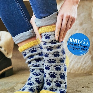 Puede incluir: Un par de calcetines de punto gris con un patrón repetitivo de huellas de patas en azul oscuro, terminados con un puño y una puntera acanalados en color amarillo mostaza. Un gráfico azul muestra el texto: KNIT for the pet lovers in your life!