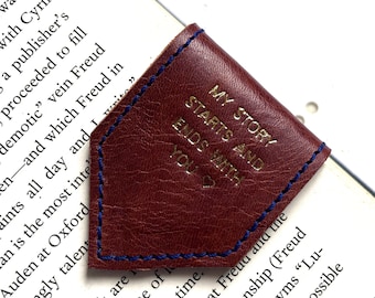 Può includere: Un segnalibro in pelle marrone con cuciture blu e lettere dorate che recitano "La mia storia inizia e finisce con te".