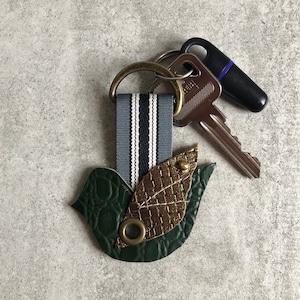 Puede incluir: Un llavero de cuero verde con forma de pájaro con un acento de hoja dorada y una cinta a rayas. El llavero está unido a un anillo de metal con dos llaves.