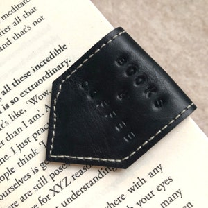 May include: A black leather bookmark with white stitching, embossed with "BOOKS CAFE". The triangular bookmark sits on a book's corner, with printed text visible. A simple, elegant design.