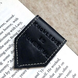 May include: A black leather bookmark with the embossed words "KNOWLEDGE IS POWERFUL" and white stitching. The bookmark is on the corner of a book, with the pages visible.