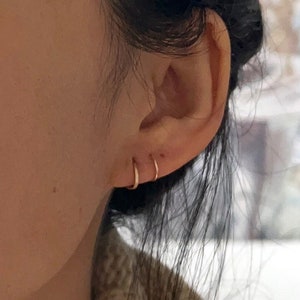 May include: Close-up of a person's ear with two small gold hoop earrings. The earrings are worn in the lobe and helix piercings.