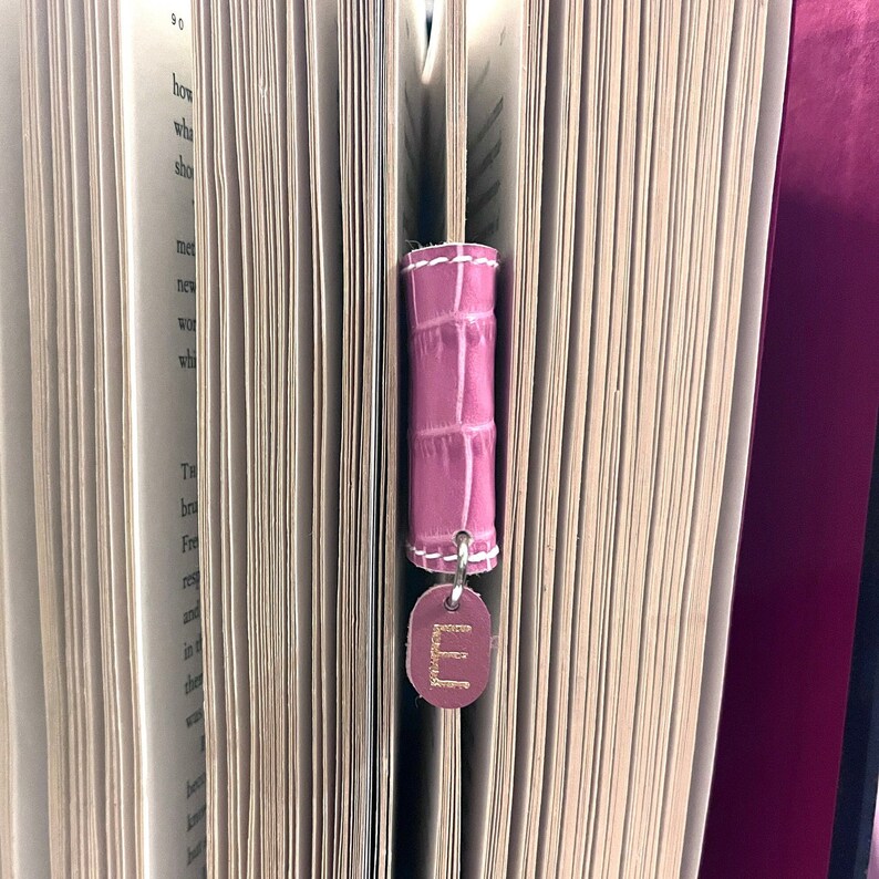 May include: A pink leather bookmark with a gold letter "E" charm. The bookmark is attached to a book with pages.