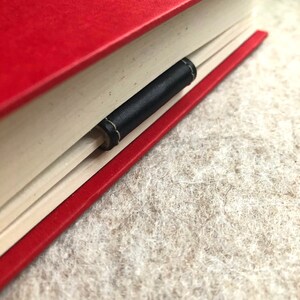 May include: A red hardback book with a black leather bookmark inserted between the pages. The bookmark is stitched with light-coloured thread. The book rests on a textured, light-coloured surface.