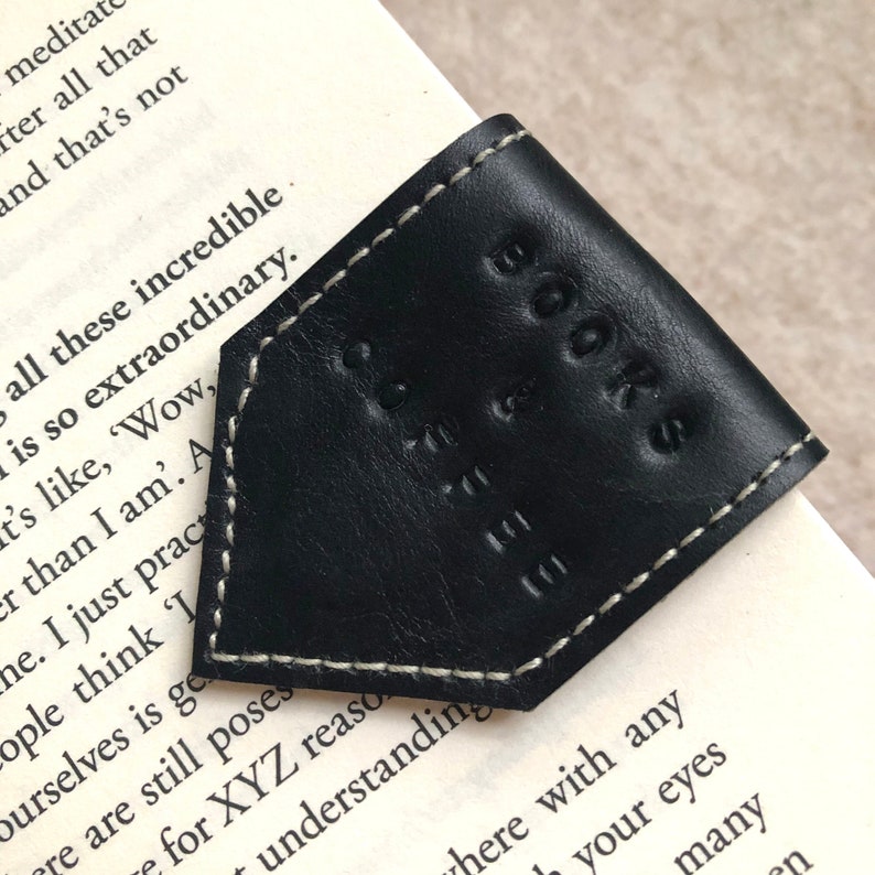 May include: A black leather bookmark with white stitching, embossed with "BOOKS CAFE". The triangular bookmark sits on a book's corner, with printed text visible. A simple, elegant design.