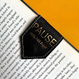 May include: Black leather bookmark with white stitching and gold lettering. The bookmark reads "PAUSE" and "HERE FOR SLEEP". The bookmark is placed on the corner of a book with printed text.