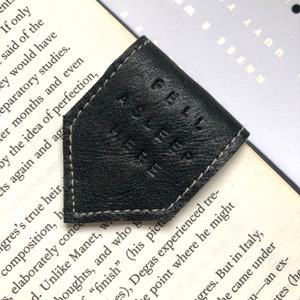 May include: Black leather bookmark with white stitching and the embossed text "A FELL ASLEEP". The diamond-shaped bookmark sits on a book's corner, a stylish accessory for any reader.