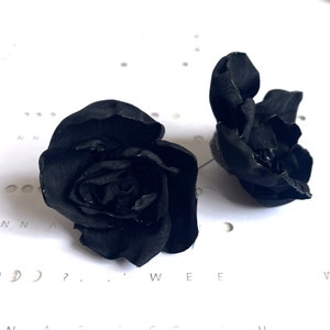 May include: Two black fabric roses, one slightly larger than the other, are shown on a white surface with a pattern of small circles.