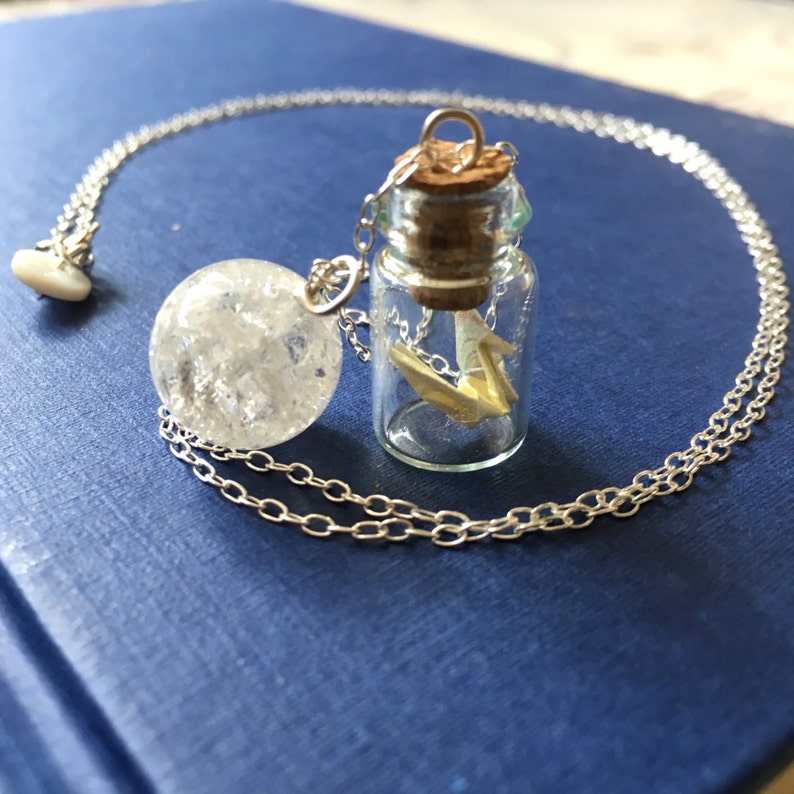 Pu&ograve; includere: Una collana a catena d'argento con un ciondolo a forma di bottiglia di vetro trasparente che contiene una barca a vela di carta bianca e un ciondolo a forma di sfera di cristallo trasparente.