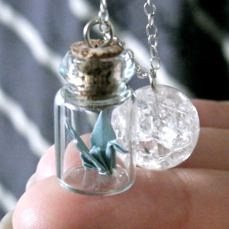 Pu&ograve; includere: Una collana delicata con un piccolo ciondolo a forma di bottiglia di vetro che contiene una gru origami blu e un ciondolo a sfera di cristallo trasparente.
