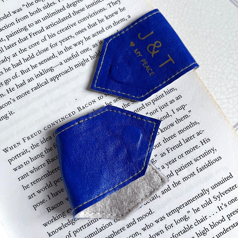 May include: Two blue leather bookmarks with white stitching. The top bookmark has the text "J&T" and "MY PEACE" in gold lettering. The bottom bookmark is plain.